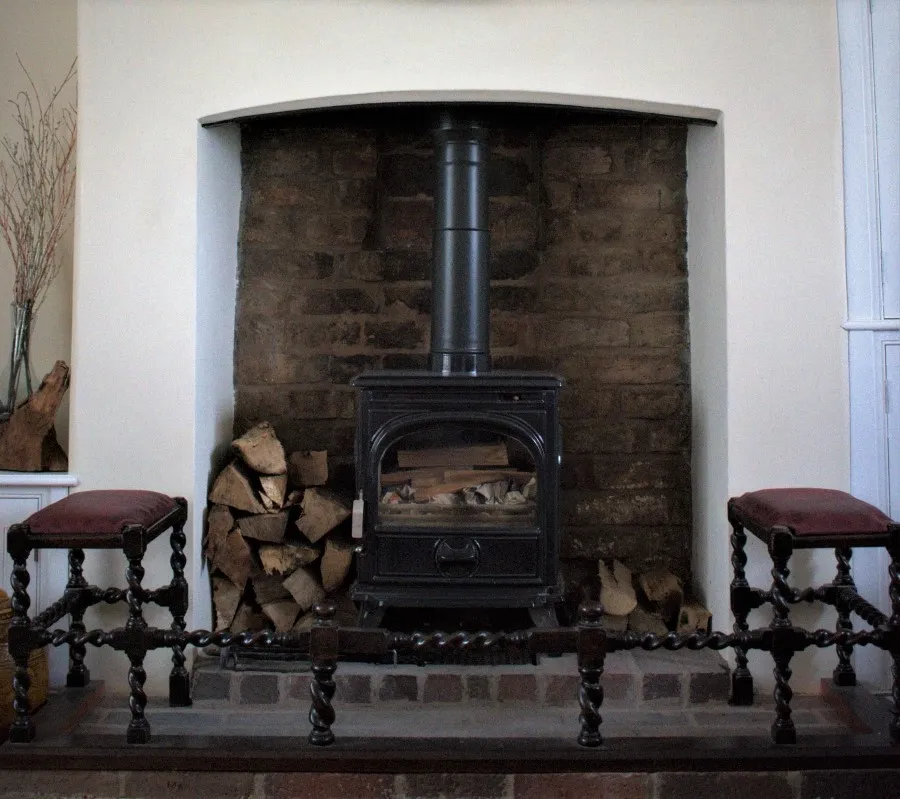 Cast iron wood stove centered in a white fireplace with stacked firewood on the left and right, and two burgundy-cushioned stools in front.