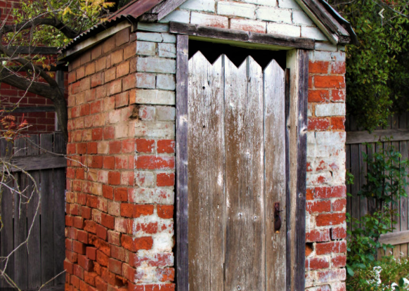 Small brick shed with a weathered wooden door and a jagged decorative trim at the top, set against a fence.Paraphrased 1