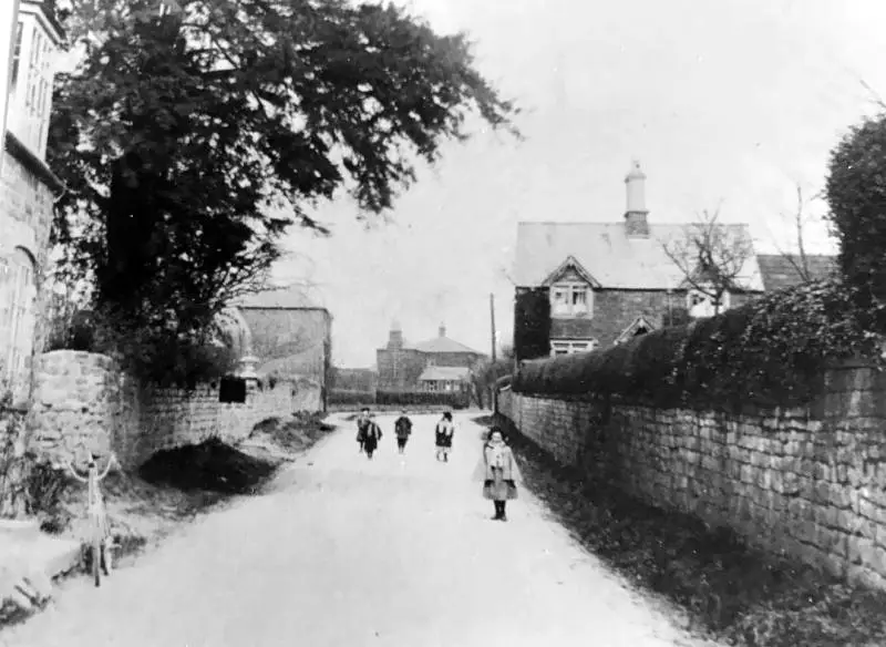 Black-and-white photo of children walking down a rural village street flanked by stone walls and houses at dusk/dawn-like light. A girl in a coat stands closer to the camera on the right side.