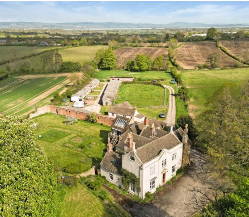 Aerial view of a white manor with chimneys, surrounded by farm buildings and green countryside fields.