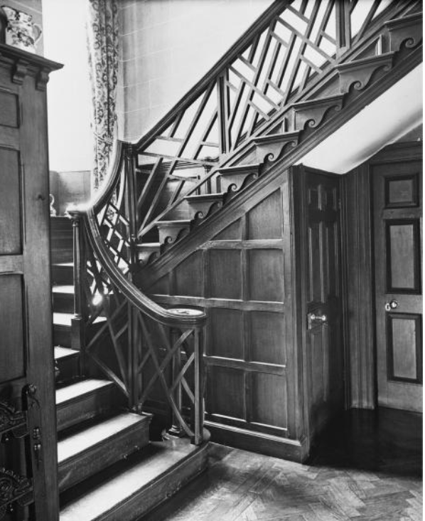 Wooden spiral staircase with carved balustrade and lattice panels, curving to an upper landing beside paneled walls and a closed door.