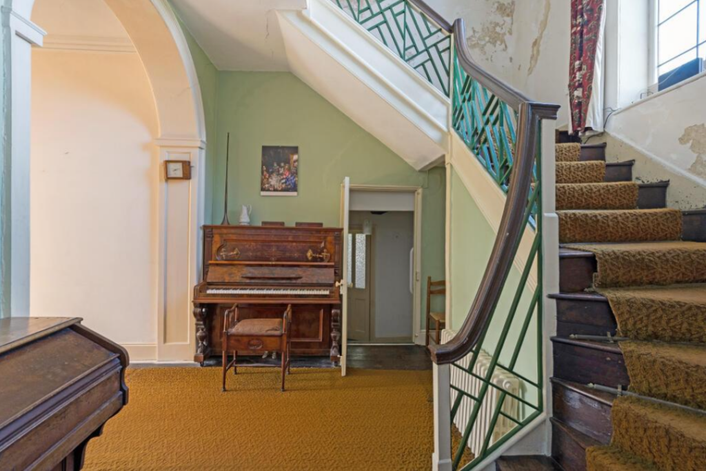 Historic interior with a curved staircase, green railing, and a wooden upright piano against a green wall under a doorway; mustard carpet.