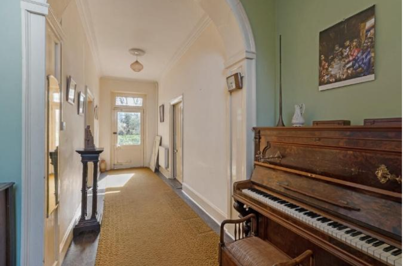 A long hallway with an arched doorway, sunlight at the far end, and a wooden upright piano against a green wall on the right.
