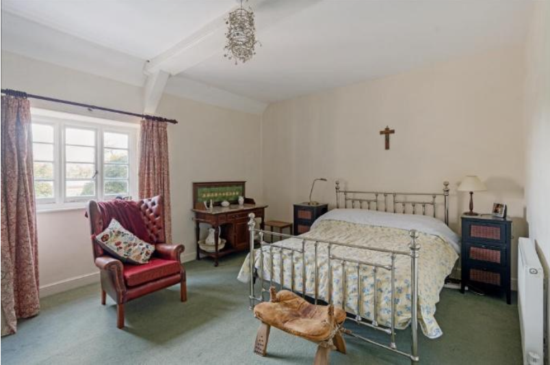 Traditional bedroom with a metal-framed bed, red armchair, and wooden dresser under a small crucifix on the wall.
