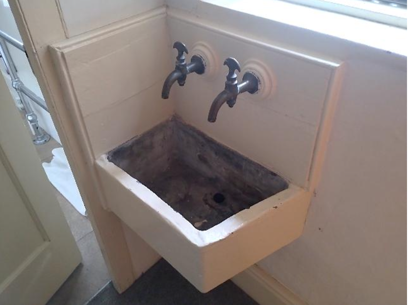White wall-mounted utility sink with two vintage taps above an open, rectangular basin showing grime inside.