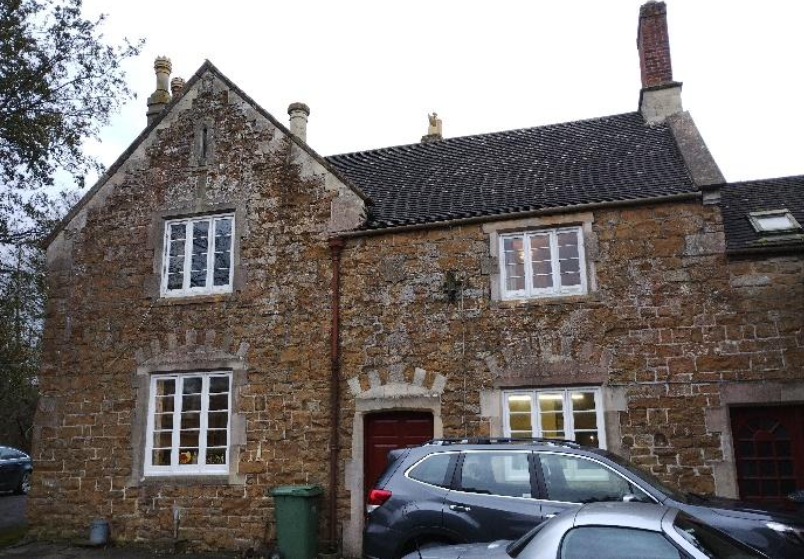 Stone cottage with white-framed windows, a red front door, and a gray car parked in front on a driveway.