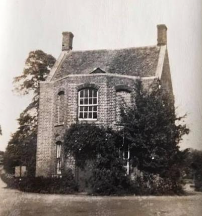 Vintage brick house with a rounded bay window, tall chimneys, and surrounding trees and bushes on a curved driveway.