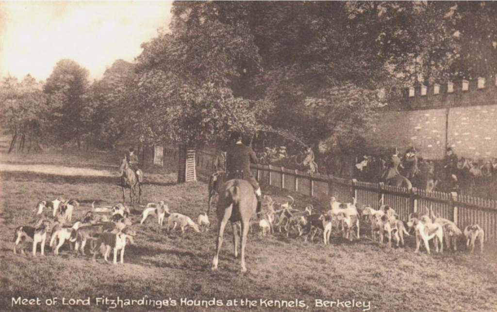 Vintage photograph of a fox-hunt meet at Berkeley kennels: riders on horses and a large pack of hounds gathered near a low fence under trees.