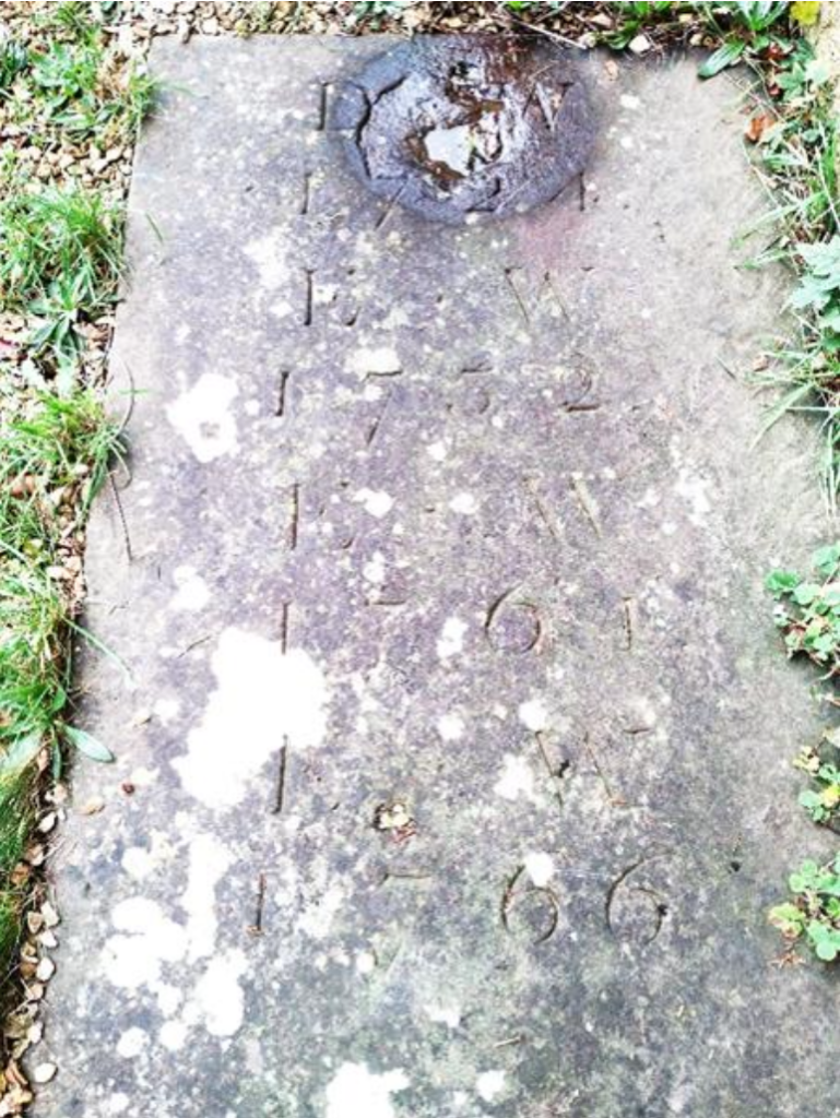 Weathered grave marker slab with faint carved numerals running down the stone, surrounded by grass and mossy patches.