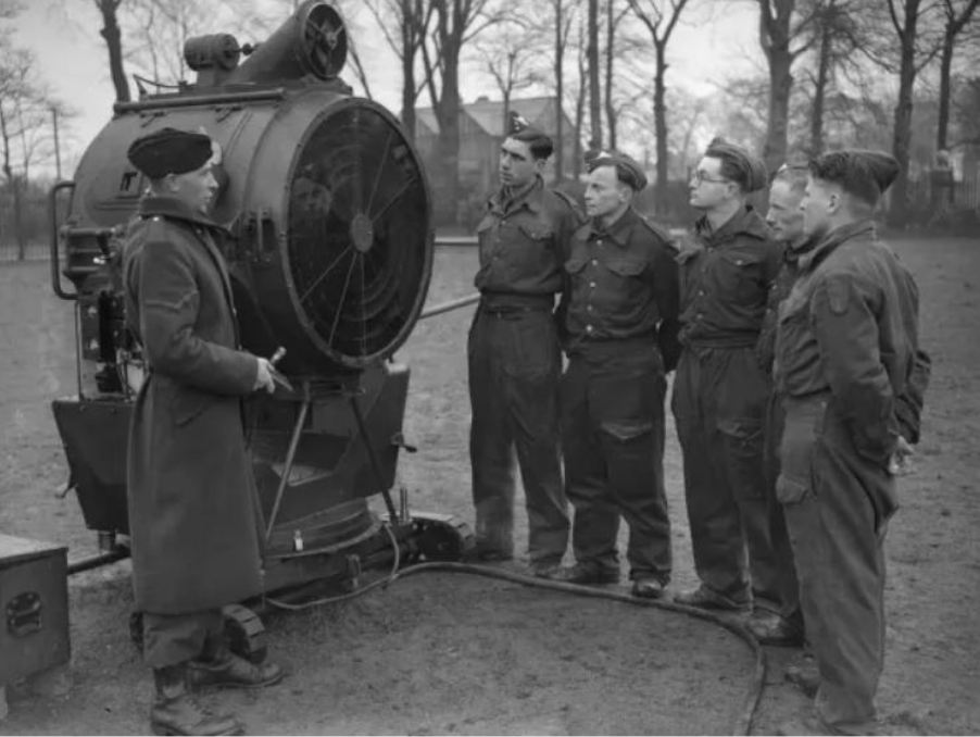 Instructor in uniform explains a large searchlight to a group of soldiers outdoors.
