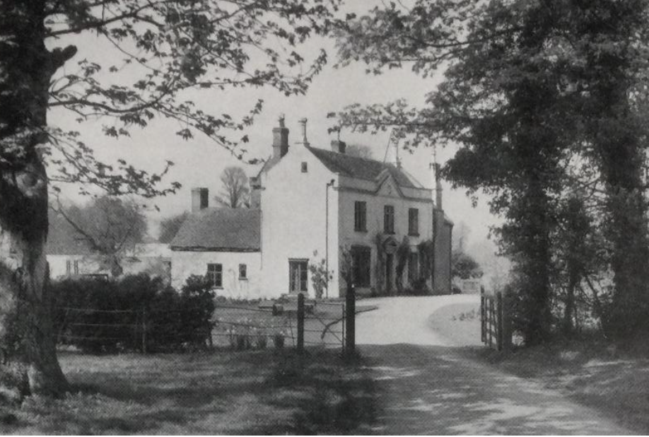 White two-story country house with a curved driveway and open wooden gate, framed by trees.