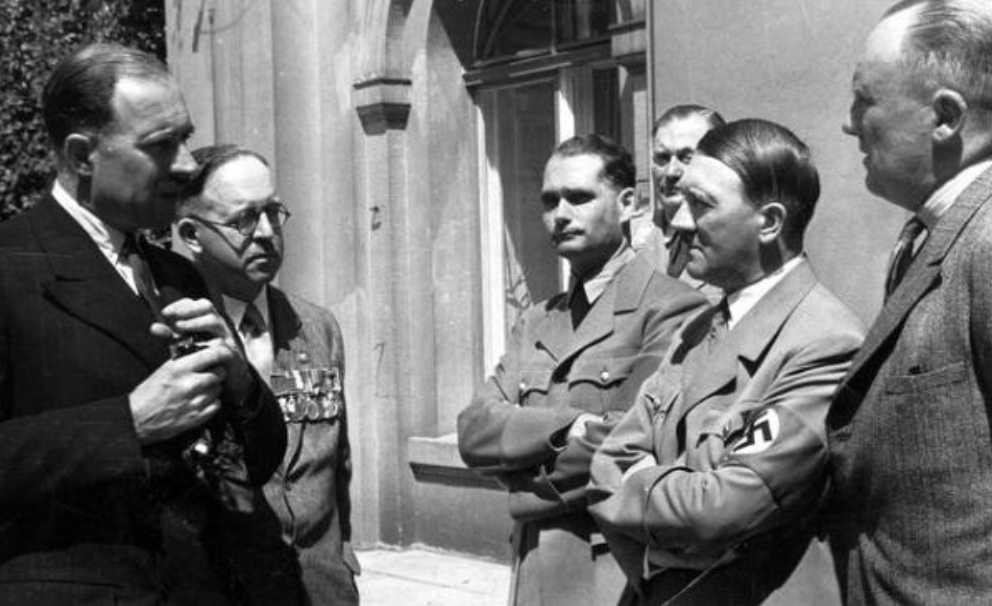 Group of men in WWII-era Nazi uniforms and suits stand outside a building, conversing; several wear medals and armbands with swastikas visible.
