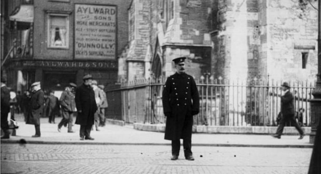 Uniformed police officer standing in the center of a cobbled street, with pedestrians and historic buildings behind.