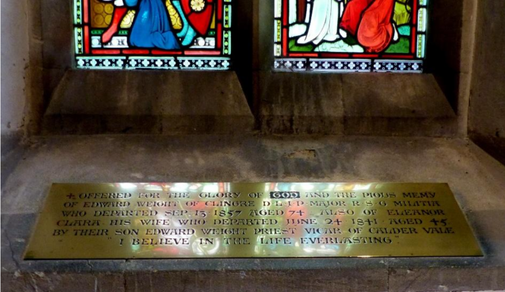 Brass memorial plaque mounted on a stone wall under two stained-glass windows, commemorating a deceased family.