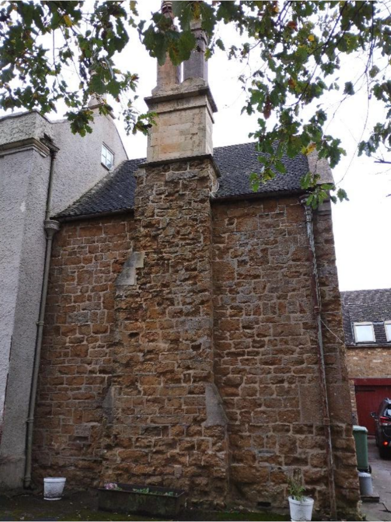 Historic stone building with a tall chimney rising from the roof, set against a leafy overhang and a neighboring modern wall on the left.