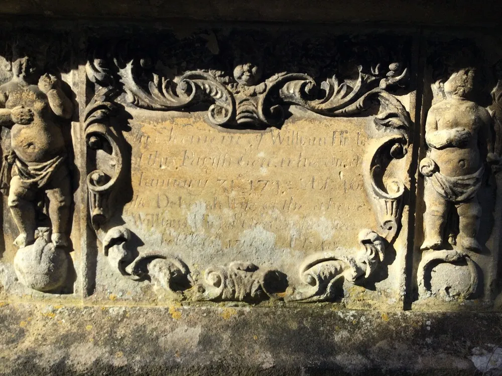 Worn stone plaque with decorative scrollwork, flanked by two carved standing figures on either side.