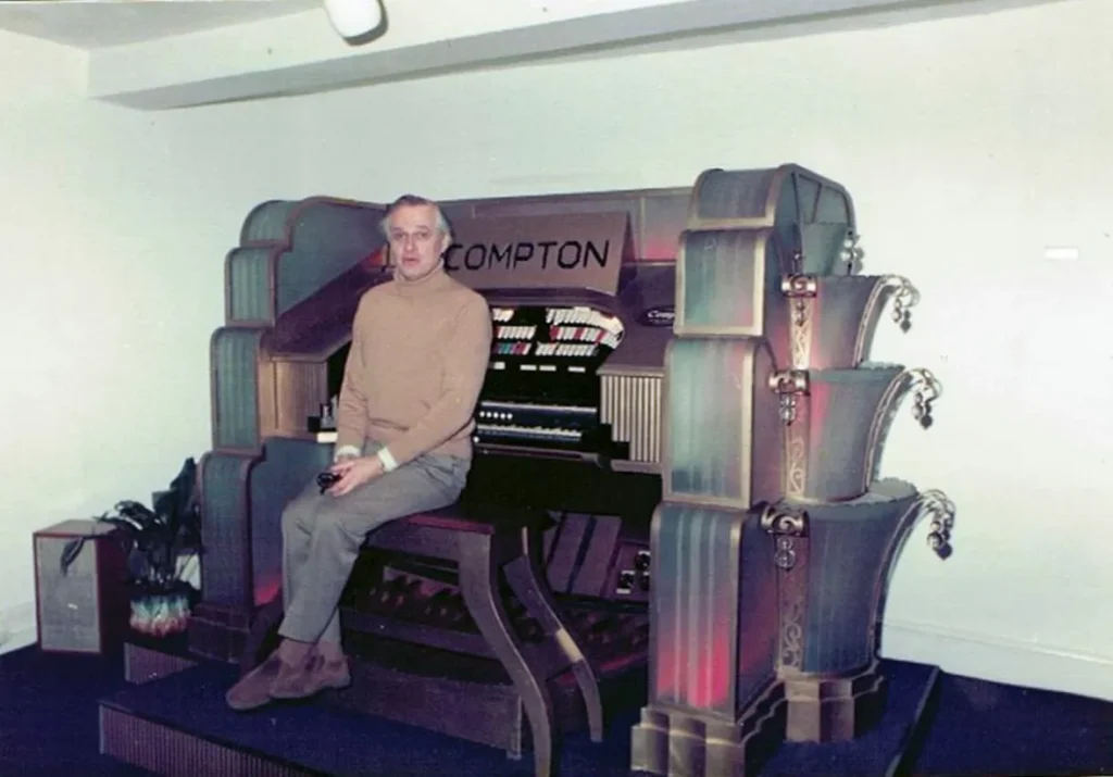 Man sitting on a vintage Compton organ with decorative side panels in a gallery-like room.