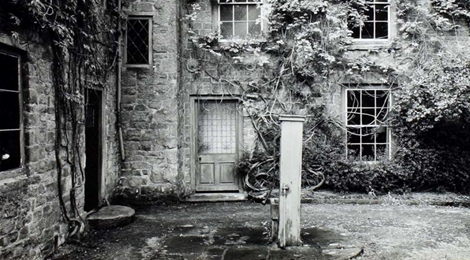 Black-and-white courtyard of an ivy-covered stone building, with a weathered wooden door, lattice windows, and a central post.
