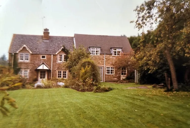 Brick two-story house with dormer windows, a sloped roof, and a small front porch, set in a well-kept lawn with shrubs and trees on a sunny day.
