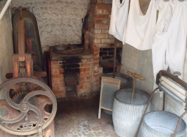 Old-fashioned laundry area with a brick oven/kiln, laundry drying line, and vintage washing tools like a hand-cranked wringer and large ceramic churns.