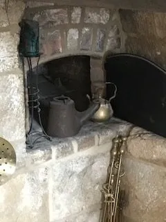 Stone fireplace with two kettles on the hearth and a brass instrument leaning against the brick surround, with a lantern nearby.