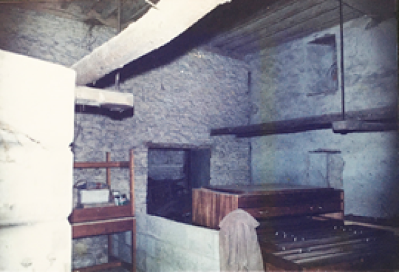 Dim stone basement room with exposed wooden beams, a large stone hearth, and simple wooden furniture along the walls.