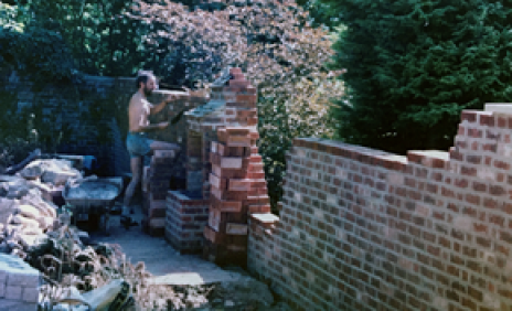Man building a brick garden wall outdoors, with stacks of bricks and a wheelbarrow nearby.