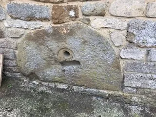Weathered stone wall with a large flat stone slab featuring a small circular hole and a short rectangular notch, likely an old water spout or trough.