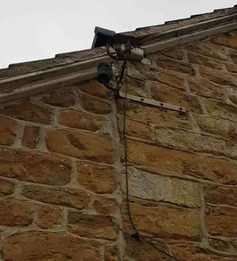 Outdoor security camera mounted on a stone wall under the roof edge, with a dangling cable down the wall.