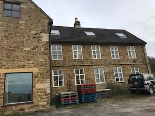 Stone two-story building with multiple windows, pallets stacked outside, and a dark SUV parked on a gravel area in front.